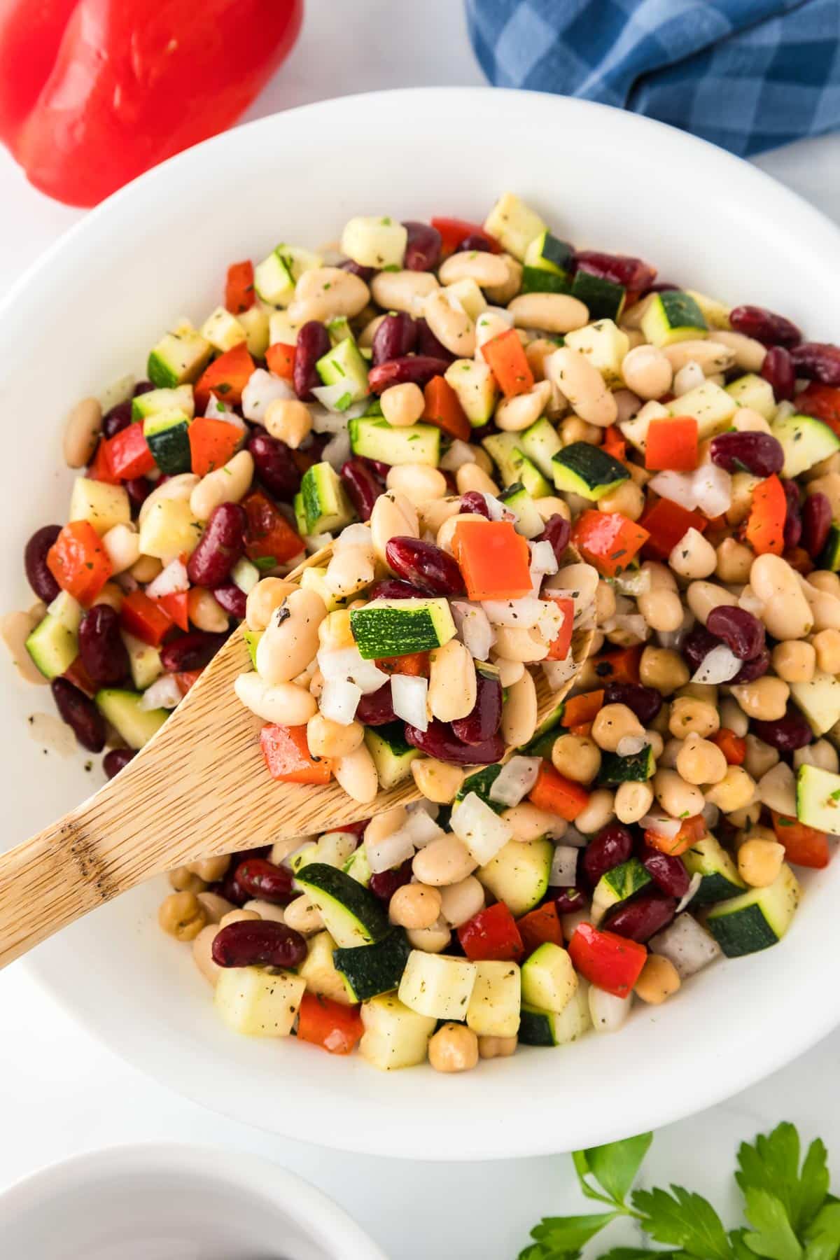 A bowl of mixed bean and chickpea salad with diced red peppers and zucchini with a wooden spoon lifting a spoonful.