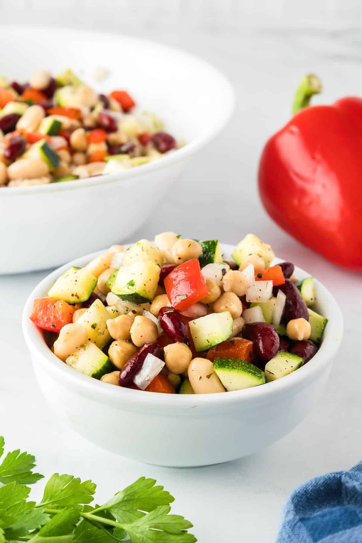 A individual bowl filled with a bean chickpea salad with a larger serving bowl full of bean salad and a bell pepper in the background on the counter.