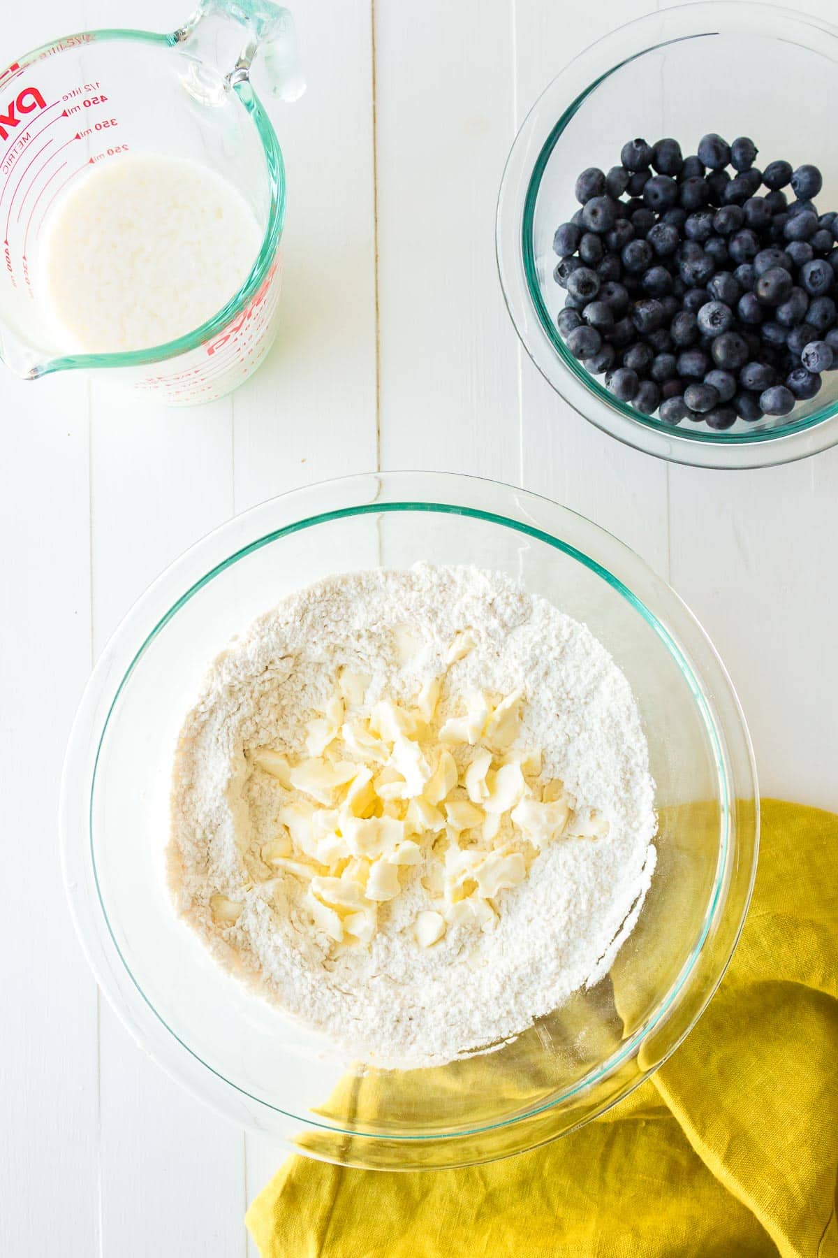 A glass bowl with a four mixture and shredded butter being mixed in with a measuring cup full of buttermilk and a bowl of blueberries nearby.