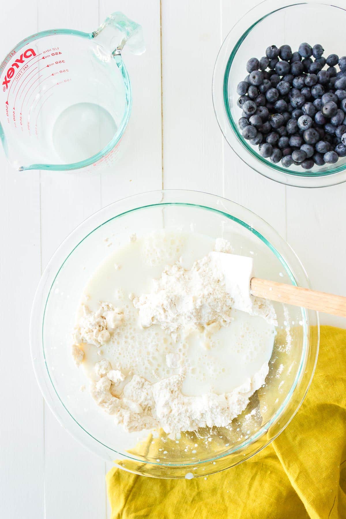 A mixing bowl with a buttermilk and flour mixture being mixed together with blueberries in a bowl nearby.