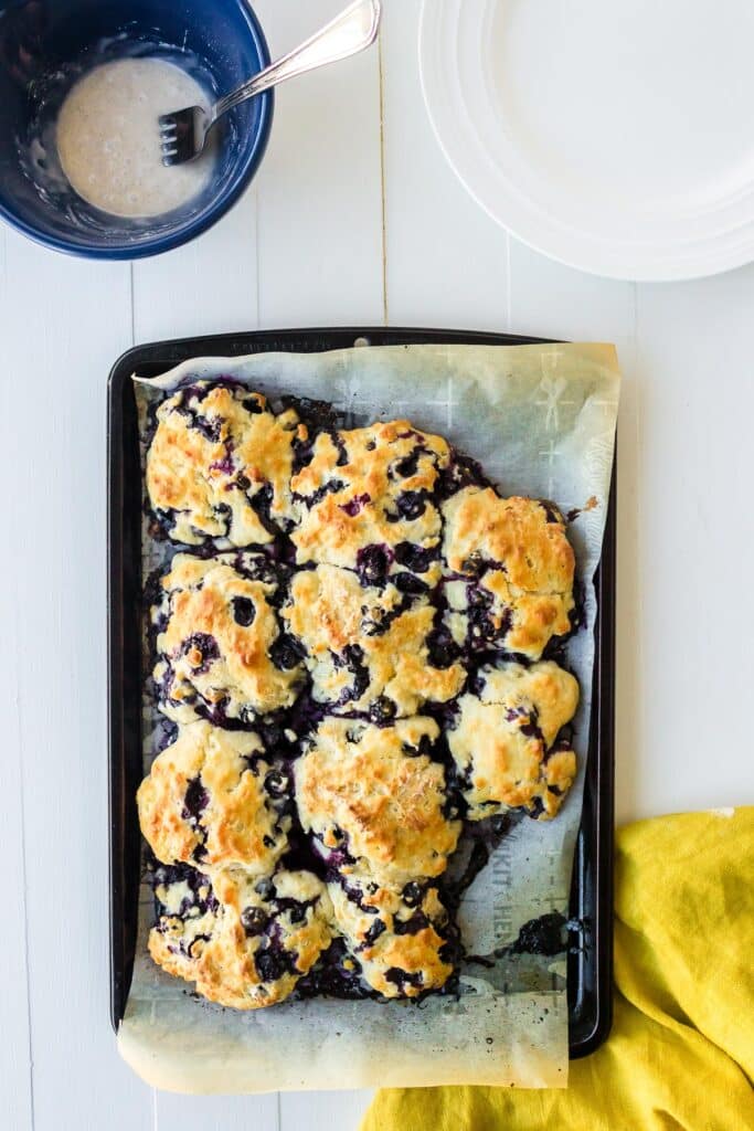 A tray of freshly baked blueberry biscuits on parchment paper with a bowl of glaze nearby.