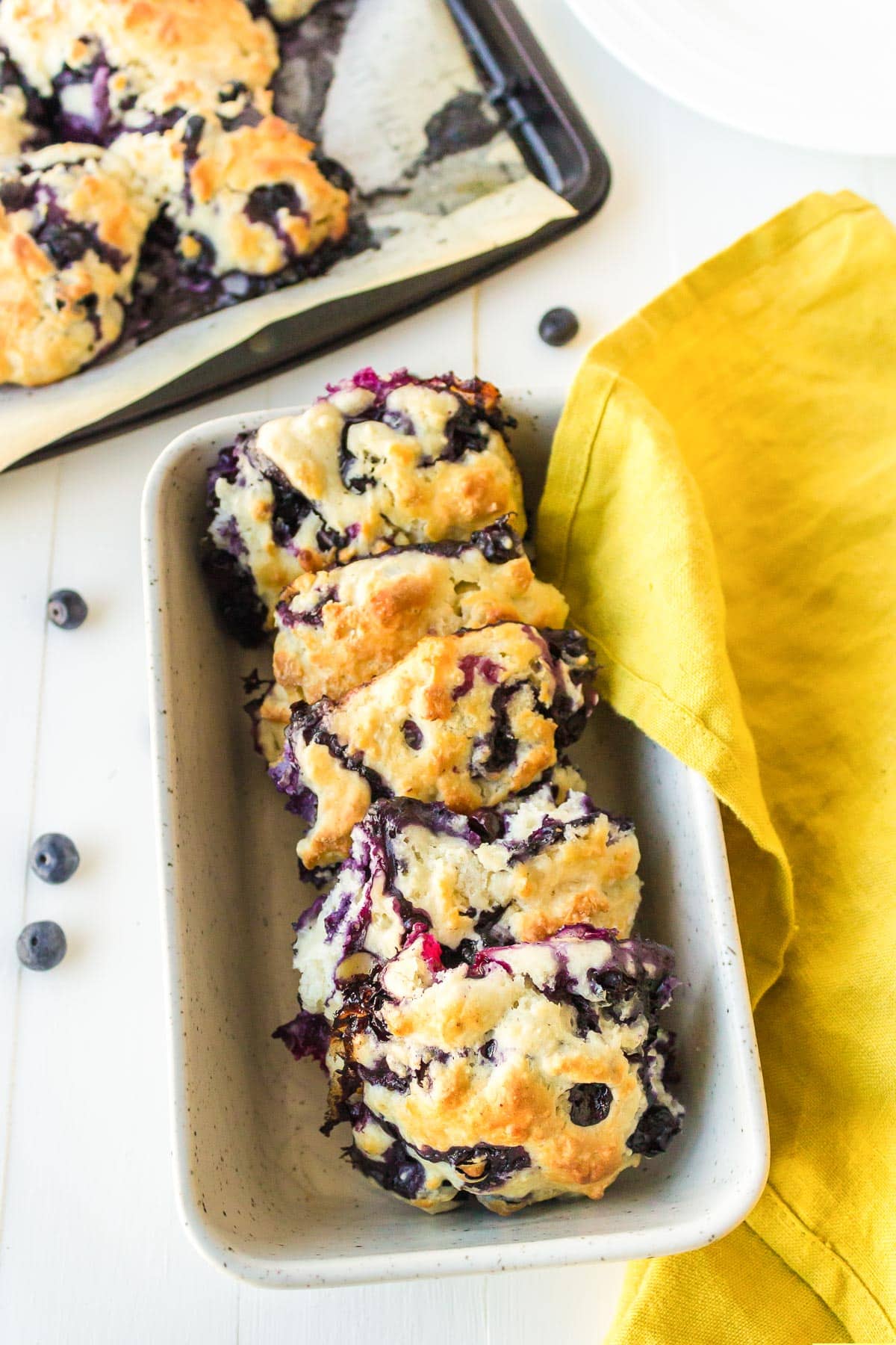 Three blueberry biscuits in a serving basket with a linen to cover them and a pan with more biscuits in the background.