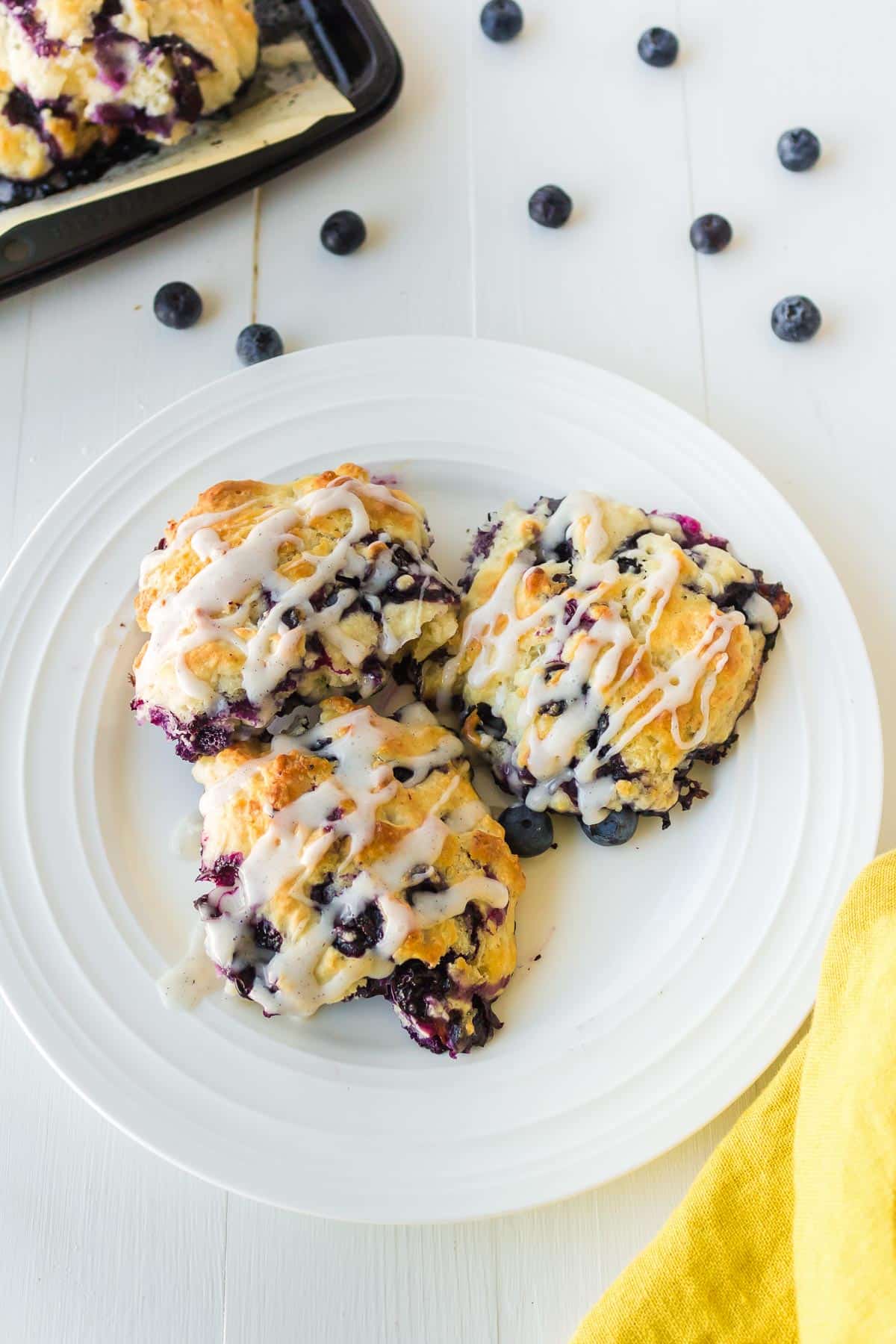 Three blueberry biscuits on a plate covered in white glaze with a pan and more blueberries scattered on the table nearby.