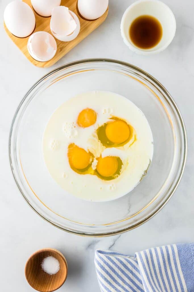 Eggs cracked in milk in a bowl next to small bowls of vanilla and salt to make the custard for brioche french toast casserole.