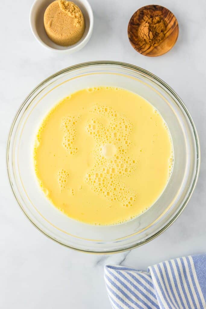 A glass bowl full of the egg and milk custard for brioche french toast after blending with brown sugar and cinnamon in small bowls nearby.