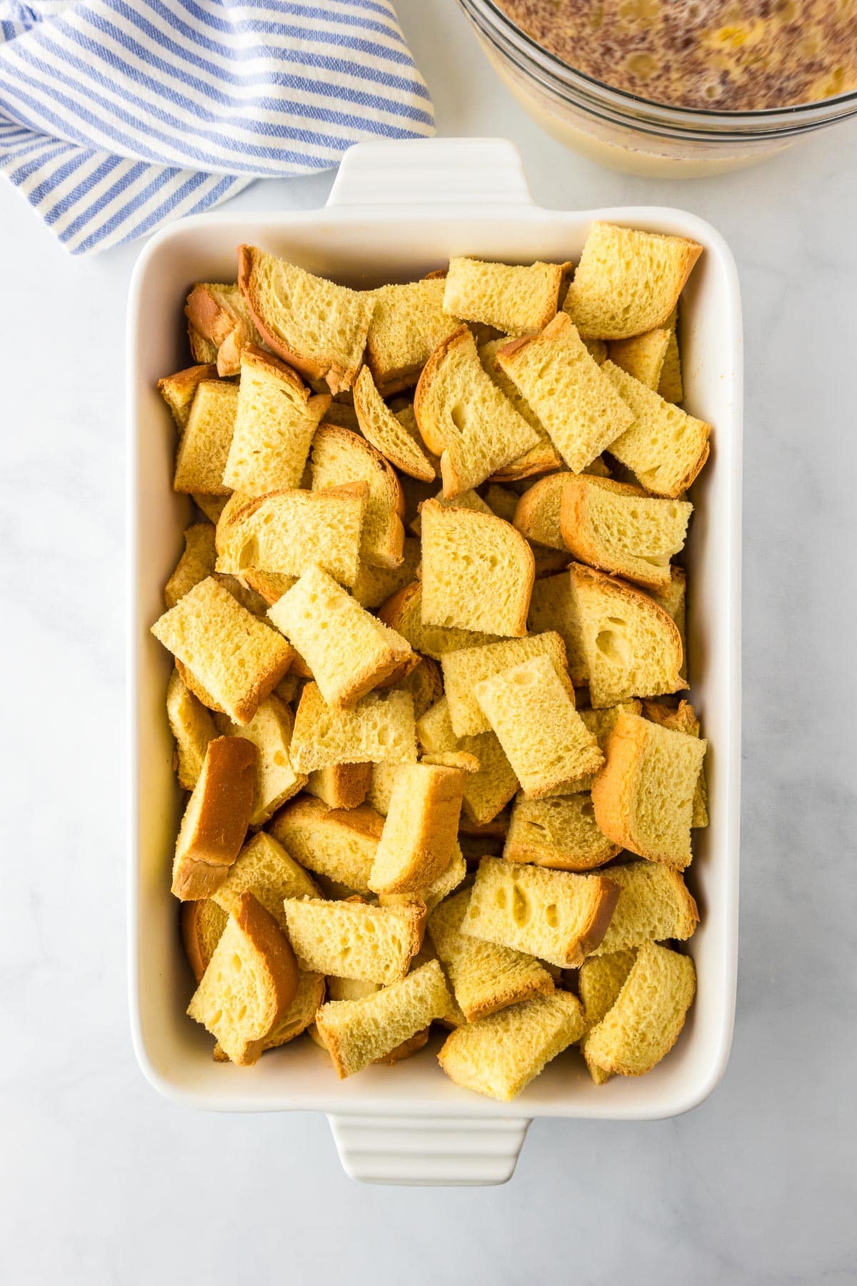 A white baking dish filled with brioche bread cubes next to a bowl full of french toast custard.