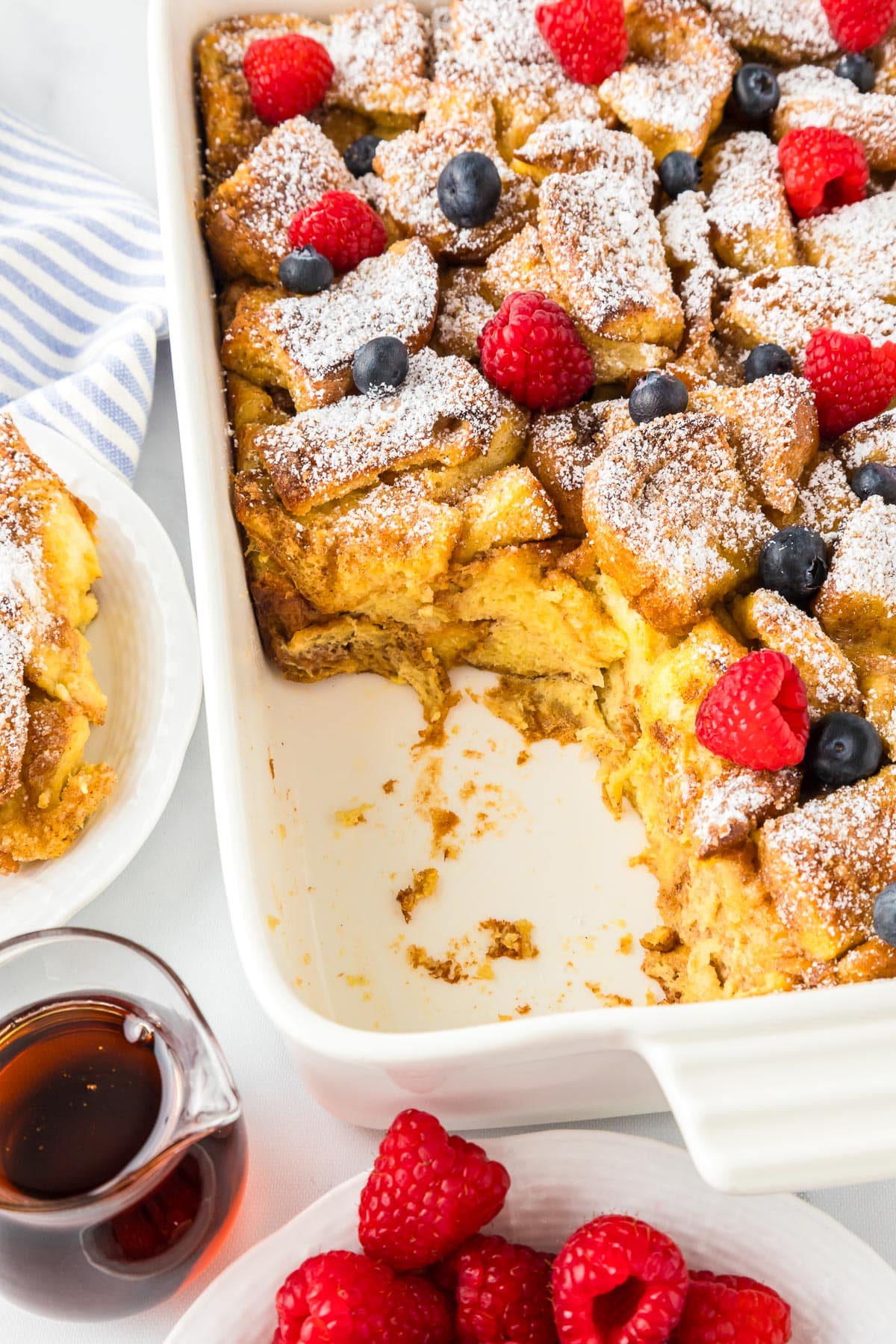 A baking dish with a partially eaten French toast casserole topped with raspberries, blueberries, and powdered sugar. A small bowl of raspberries and a jug of syrup are beside it.