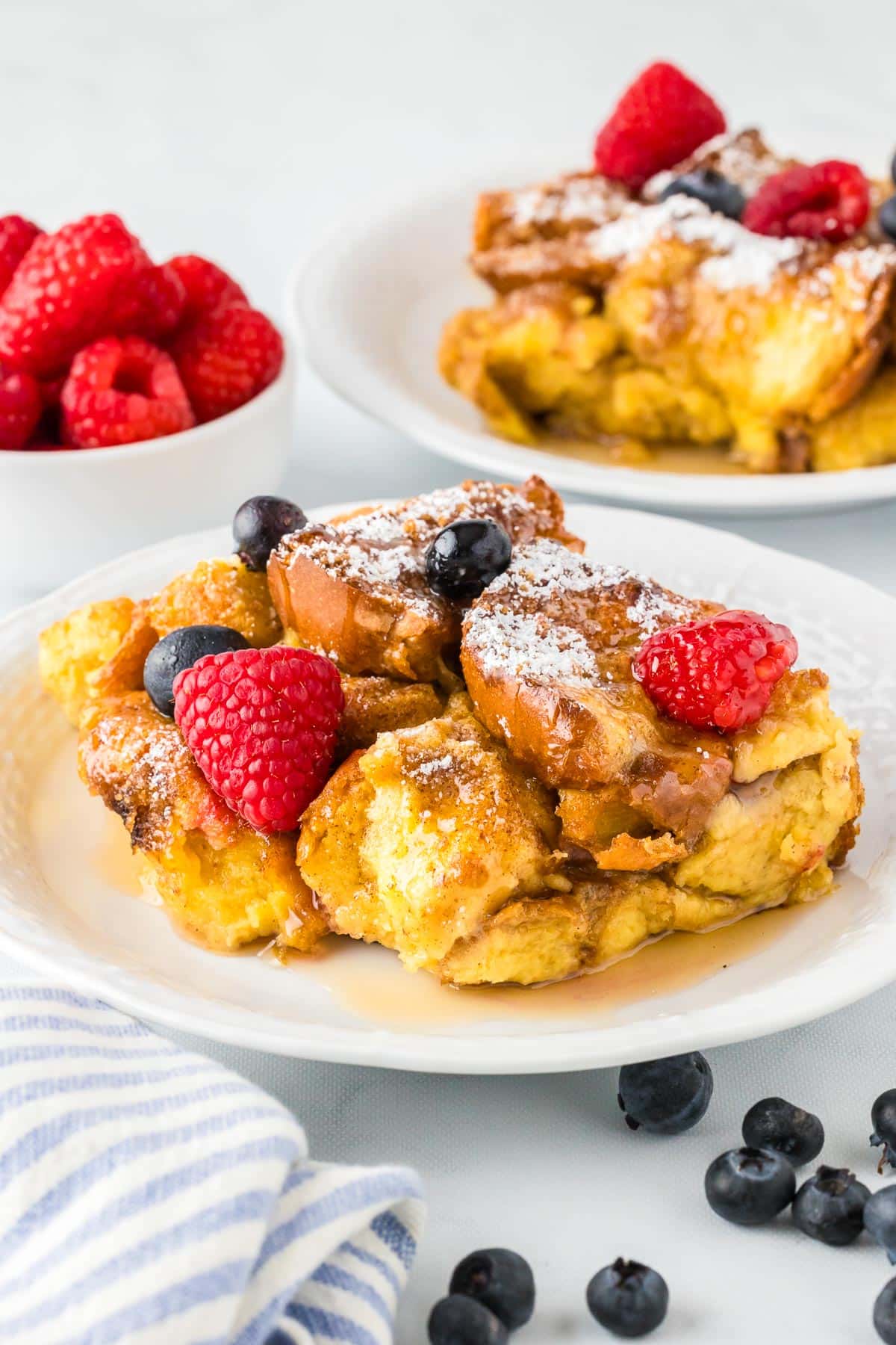 Brioche french toast casserole on a plate topped with raspberriee, blueberries and powdered sugar with more french toast on a plate and berries in the background.