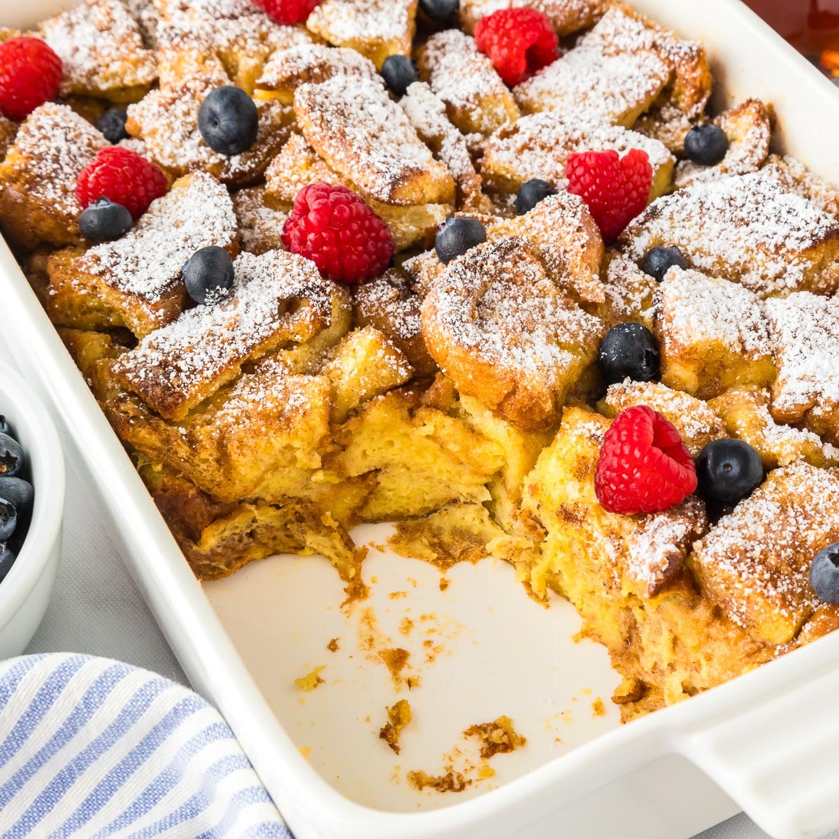 Square view of brioche French toast casserole in a baking pan missing a scoop topped with berries and powdered sugar.