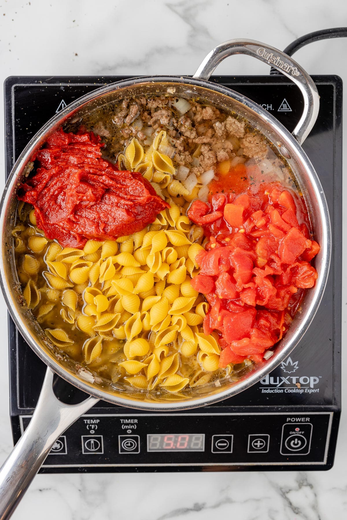 A large pan fill of cooked ground beef, diced tomatoes, tomato paste and dry shells pasta before cooking.