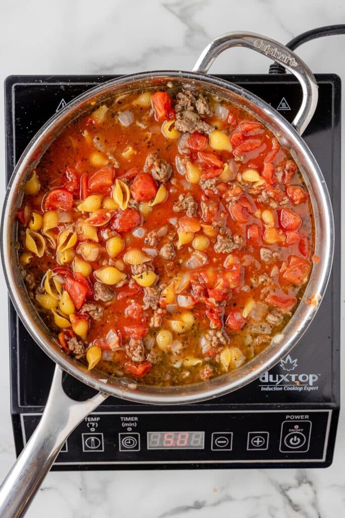 A large pan with tomatoes, ground beef and pasta shells cooking to make homemade hamburger helper on a burner.