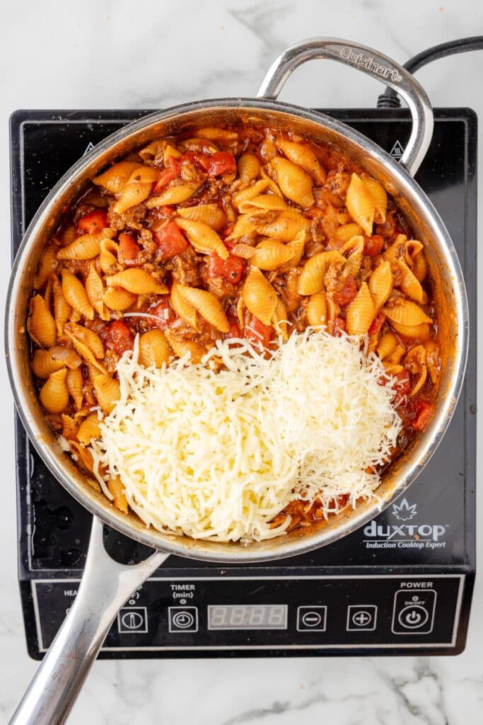 A large pan with cooked homemade hamburger helper with shells with shredded cheese being added to the pan on a burner.