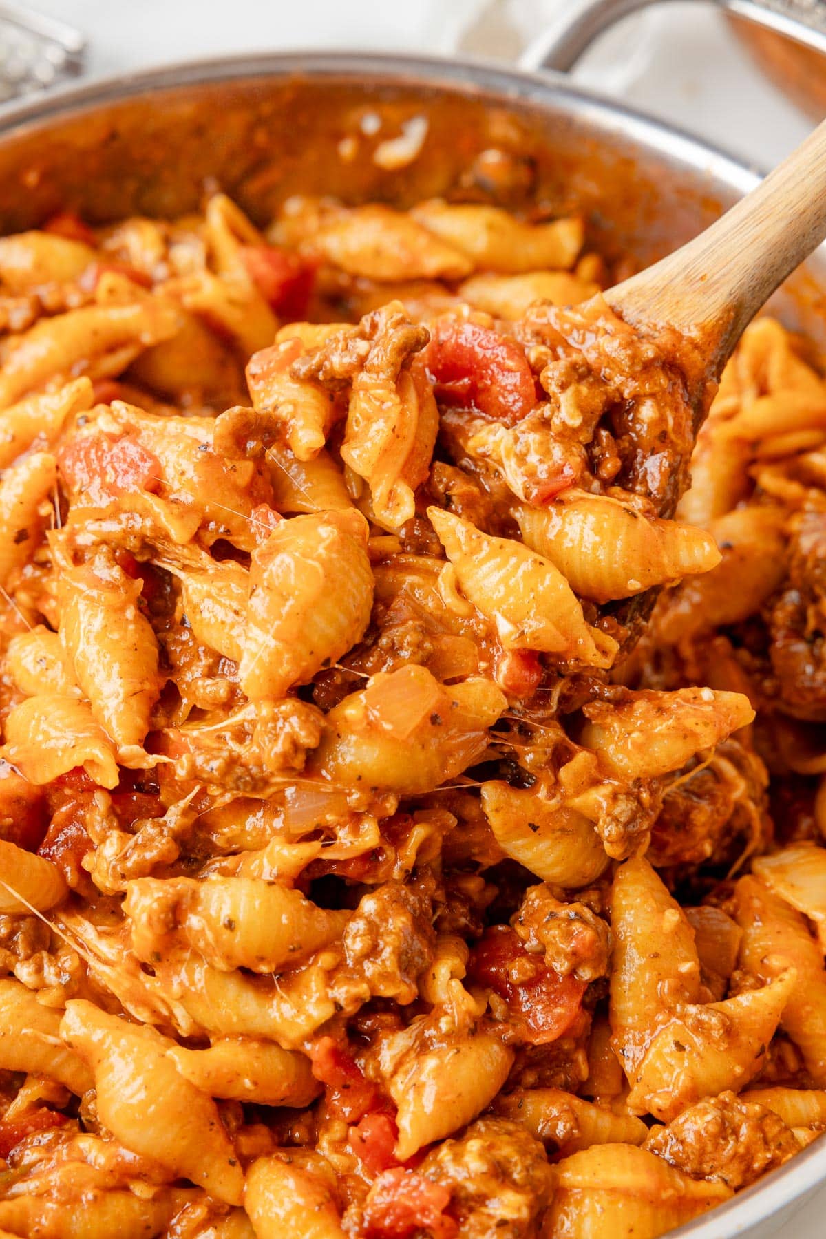 Close-up of a skillet filled with cheesy homemade hamburger helper shells being scooped by a wooden spoon.