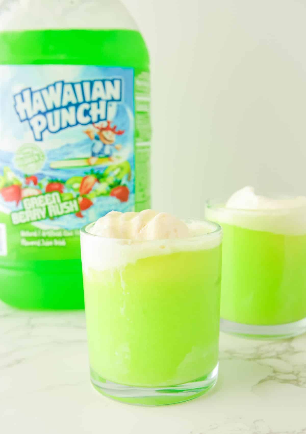 Two glasses of light green punch with ice cream floating to make a Halloween punch in front of a large bottle of Hawaiian Punch Green Berry Rush.