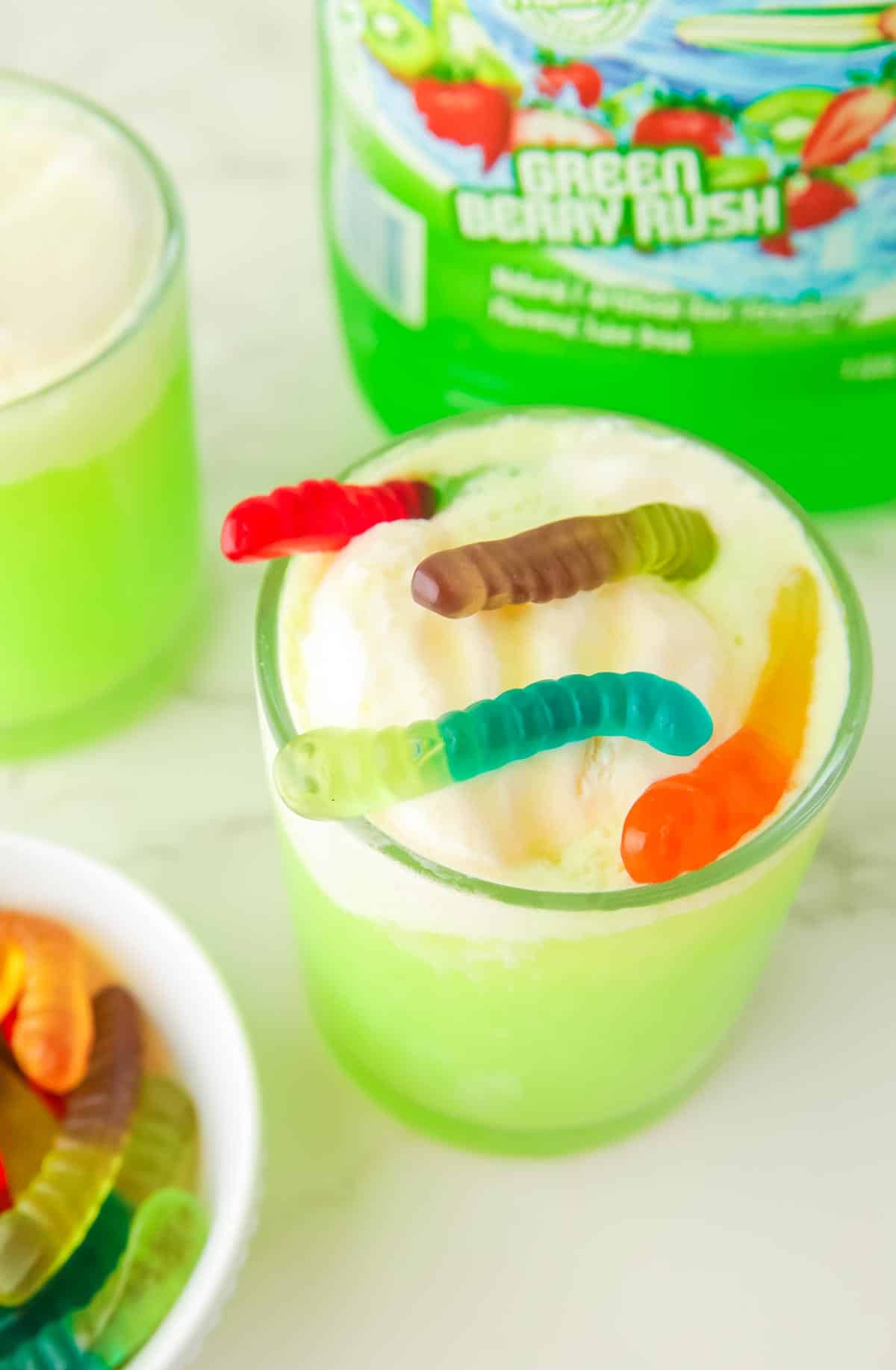 Green Halloween punch topped with ice cream and gummy worms with more punch on the counter in the background.