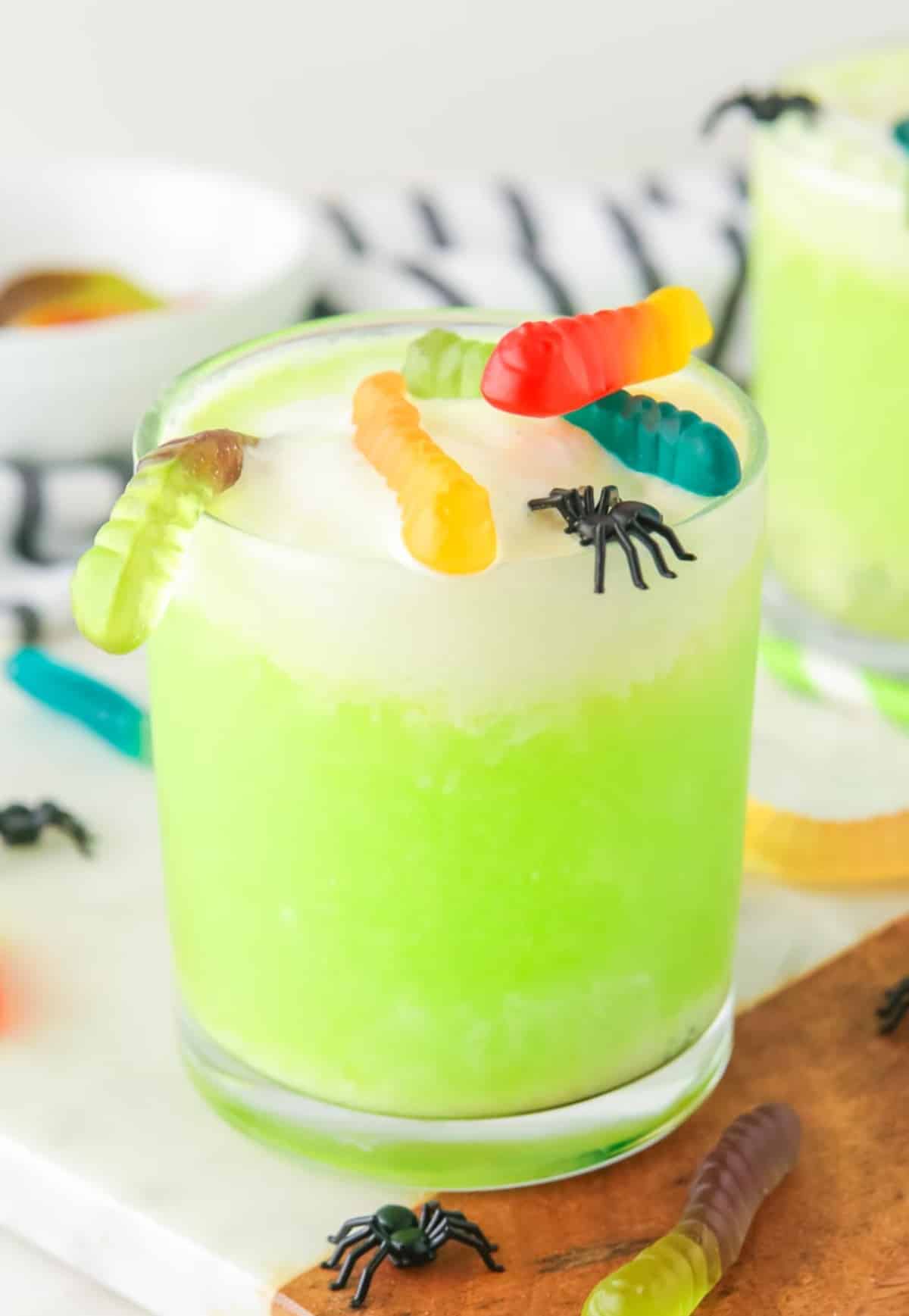 A glass of green Halloween punch topped with gummy worms and gummy bugs with a second glass in the background.