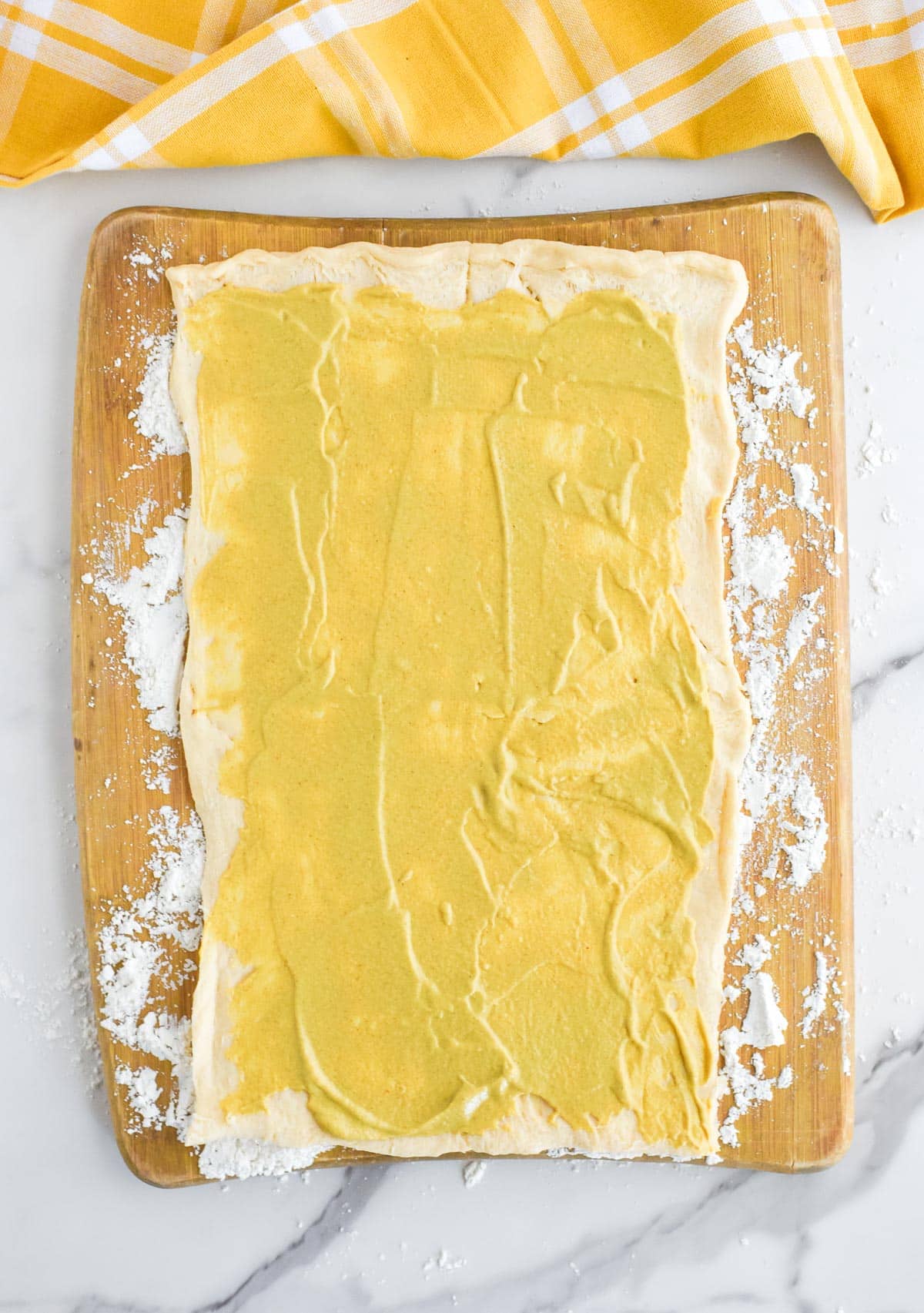 Unrolled crescent roll dough on a wooden cutting board dusted with flour with a thin layer of Dijon mustard spread over the dough.