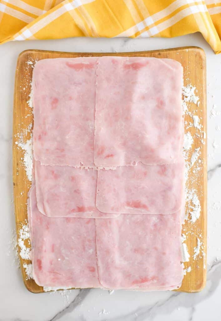 A wooden cutting board with crescent roll dough laid out and layered with ham.