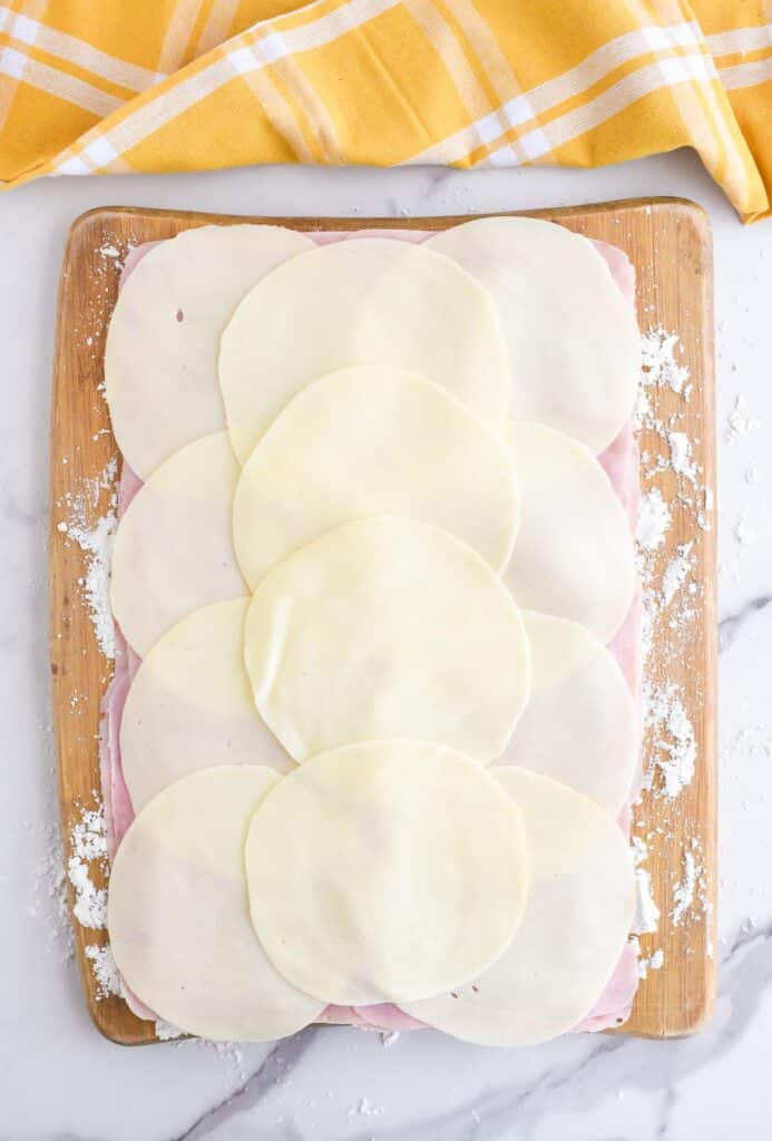 A wooden cutting board with crescent roll dough laid out and layered with provolone cheese and ham.