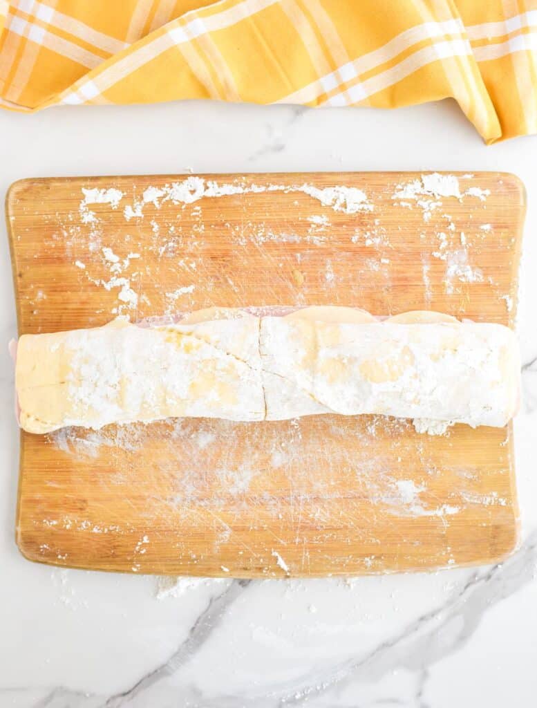 A wooden cutting board coated lightly with flour with rolled ham and cheese pinwheel dough right after rolling.