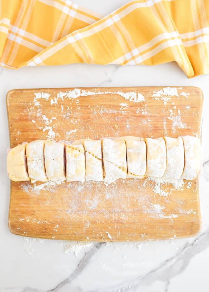 A wooden cutting board with rolled ham and cheese pinwheel dough cut into even pieces.