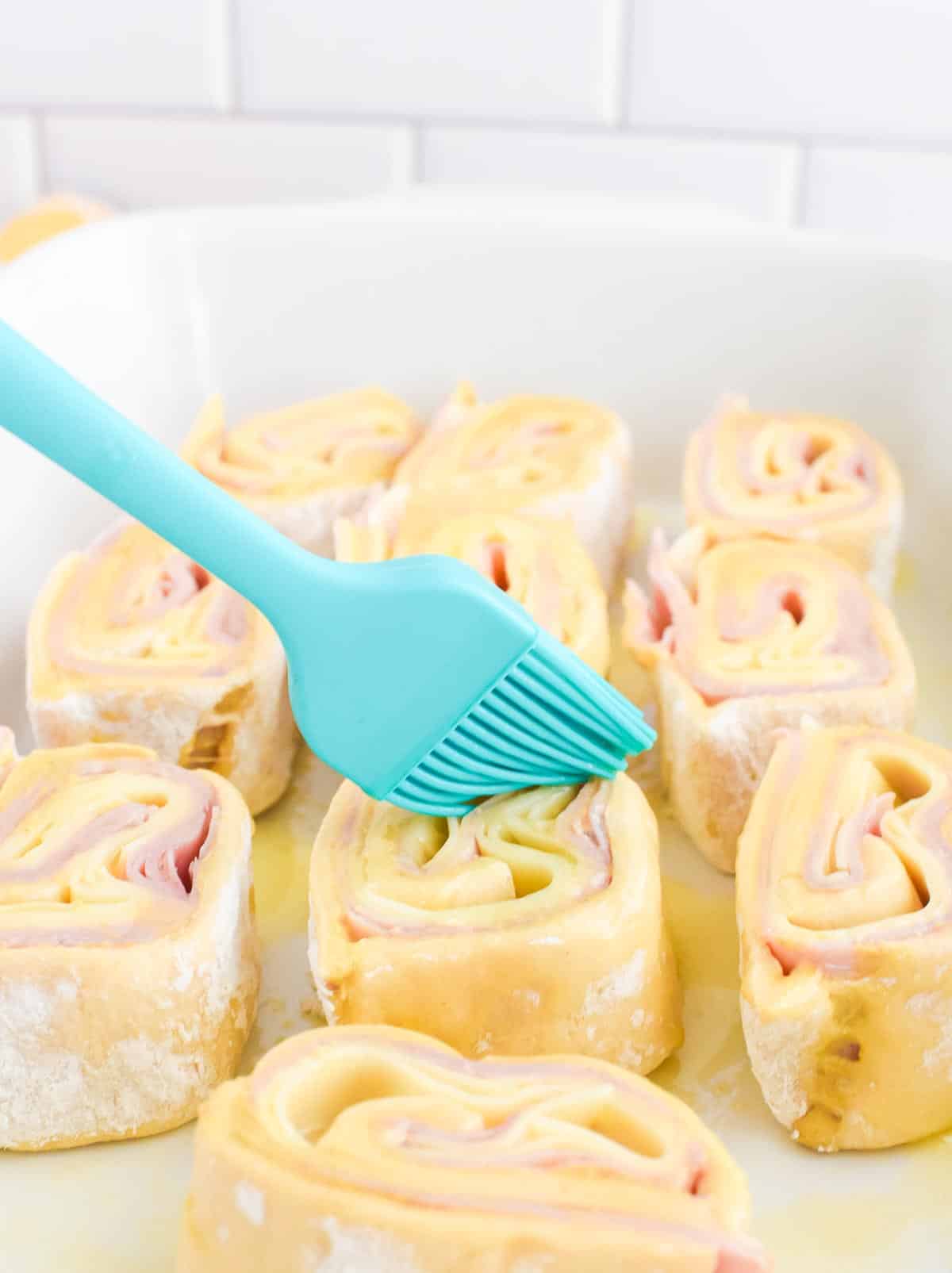 A silicone brush adding melted butter to unbaked ham and cheese pinwheels in a baking dish.