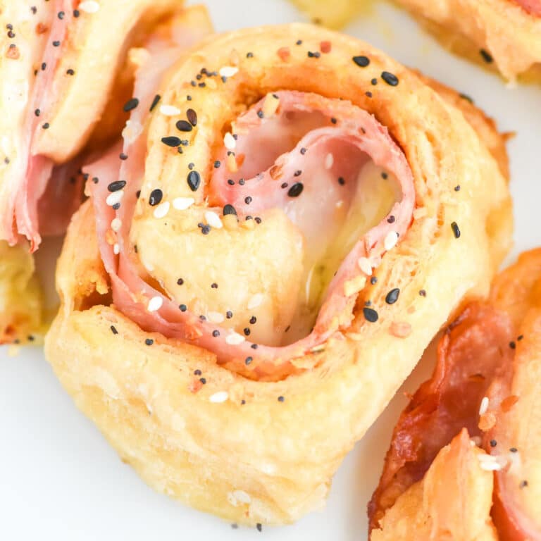 Close-up of a ham and cheese pinwheel roll with sesame seeds on top, showing layers of ham and melted cheese inside a golden-brown dough.