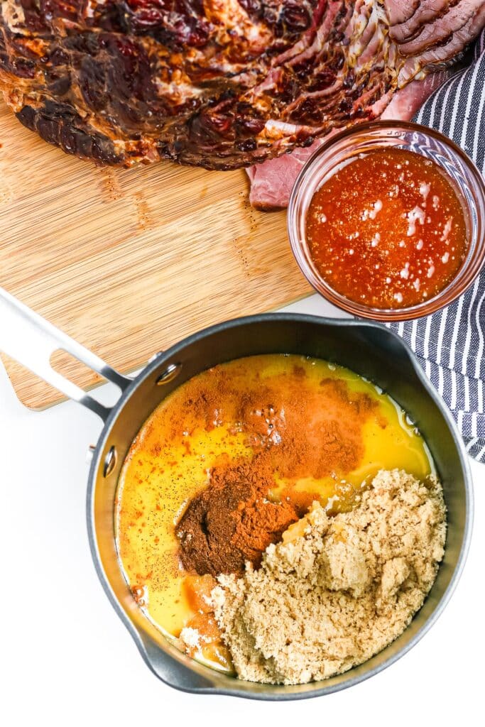 Ingredients for honey orange ham glaze being mixed in a small pan with a small bowl of honey and a full ham on a cutting board nearby.