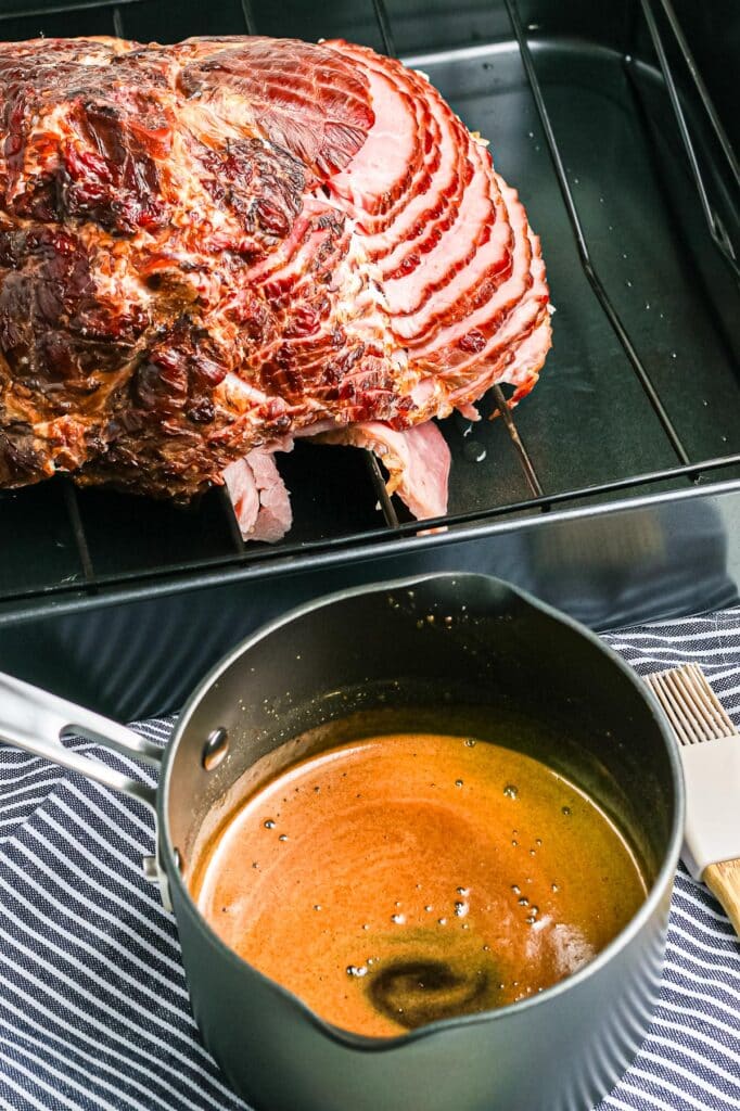 A baked ham, sliced and placed in a roasting pan next to a sauce pan with a honey orange ham glaze and a silicone brush.