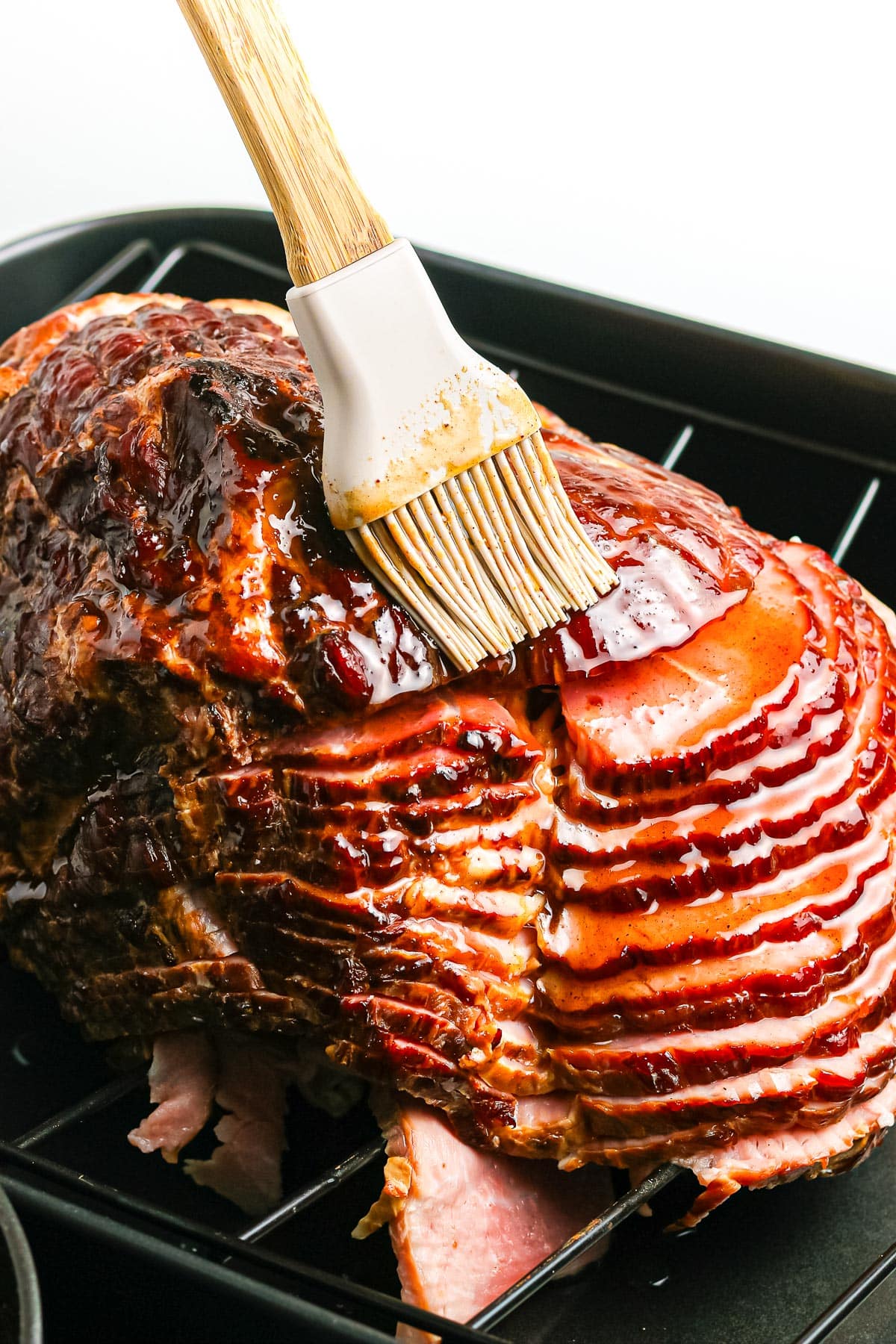 A sliced ham sitting on a roasting pan with a silicone brush glazing the ham.