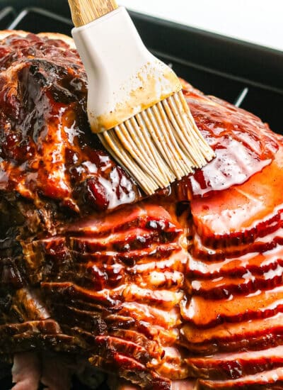 Silicone brush glazing a sliced ham in a roasting pan.