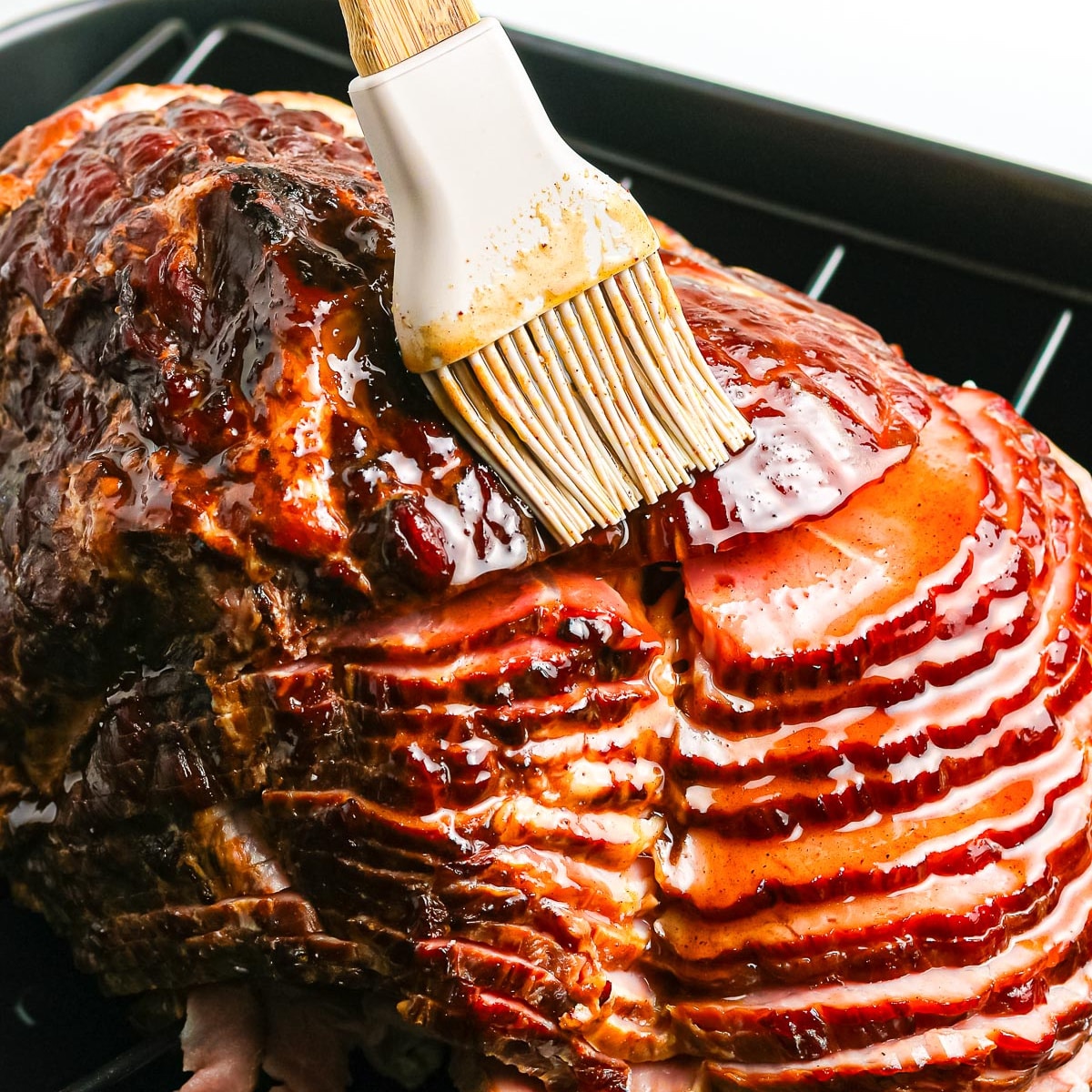 Silicone brush glazing a sliced ham in a roasting pan.