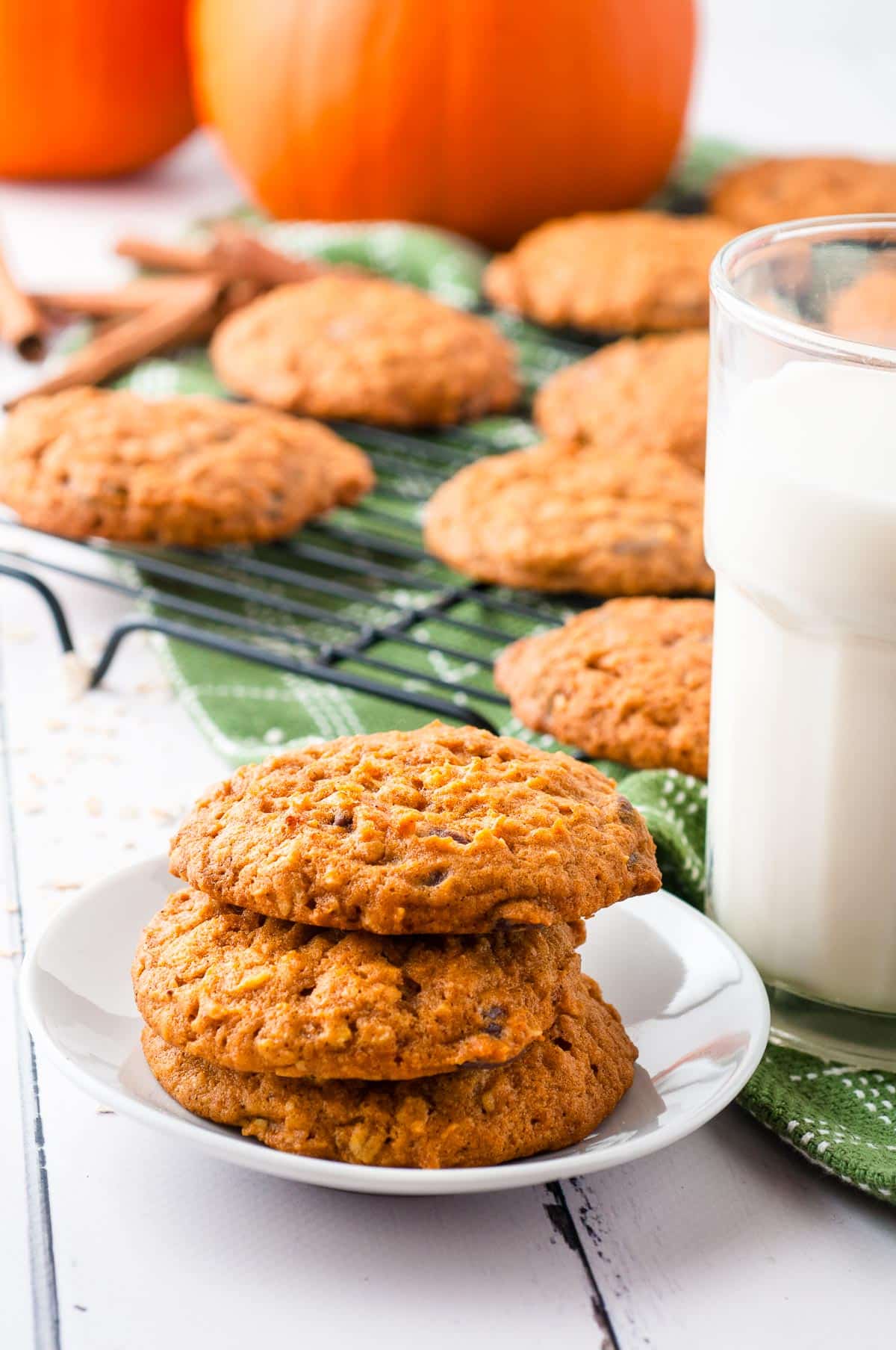 A plate with three pumpkin oatmeal cookies in front of a cooling rack full of more cookies with a glass of milk and a pumpkin in the background.