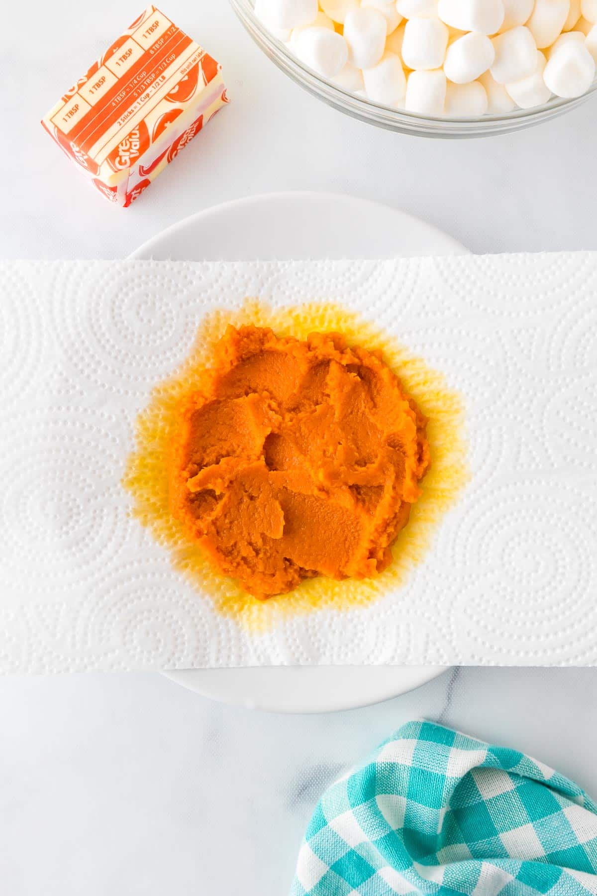 Pureed pumpkin being blotted on a paper towel to absorb excess moisture with marshmallows and butter nearby on the counter.