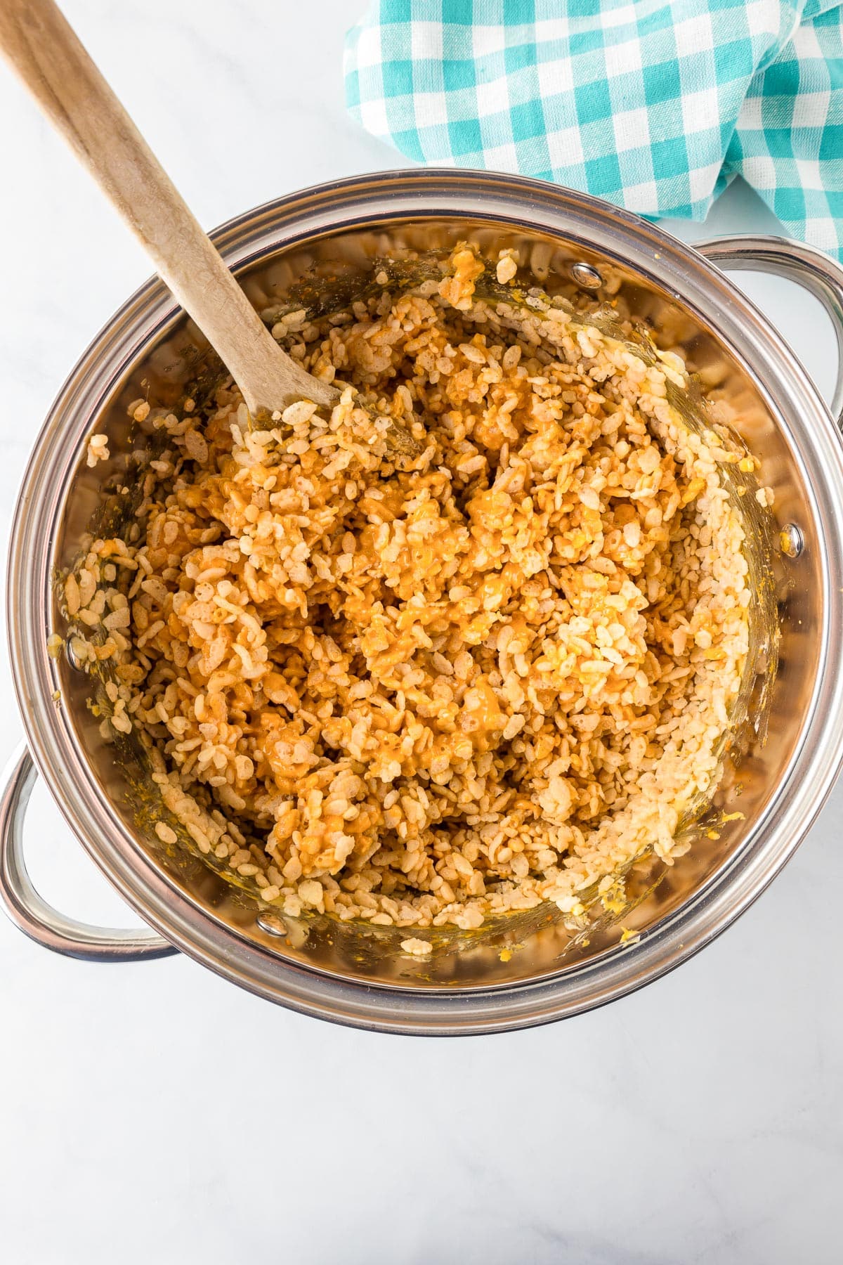 A pot filled with a mixture of orange pumpkin spice marshmallow mixture and Rice Krispies being mixed with a large wooden spoon.