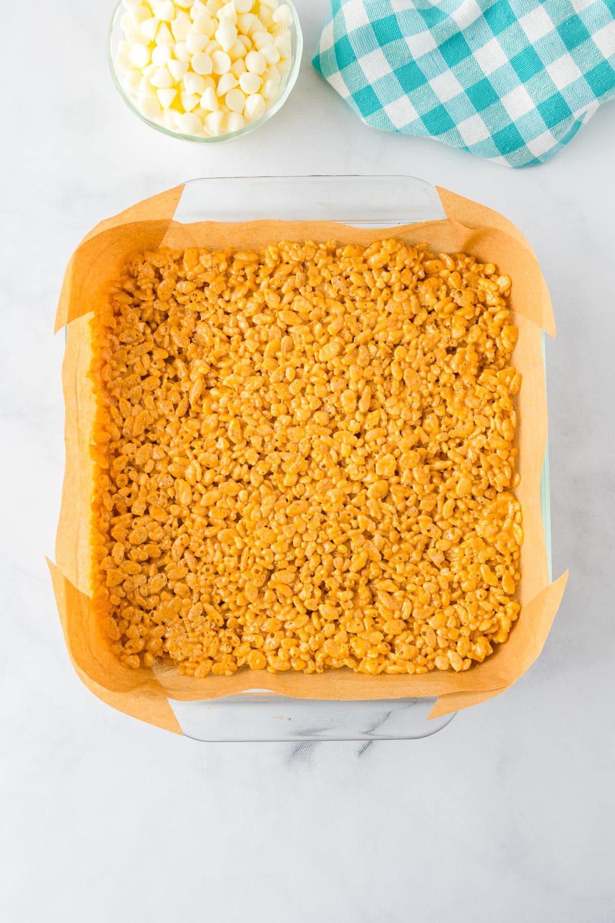 A square glass baking dish lined with parchment paper full of orange pumpkin spice Rice Krispie mixture.