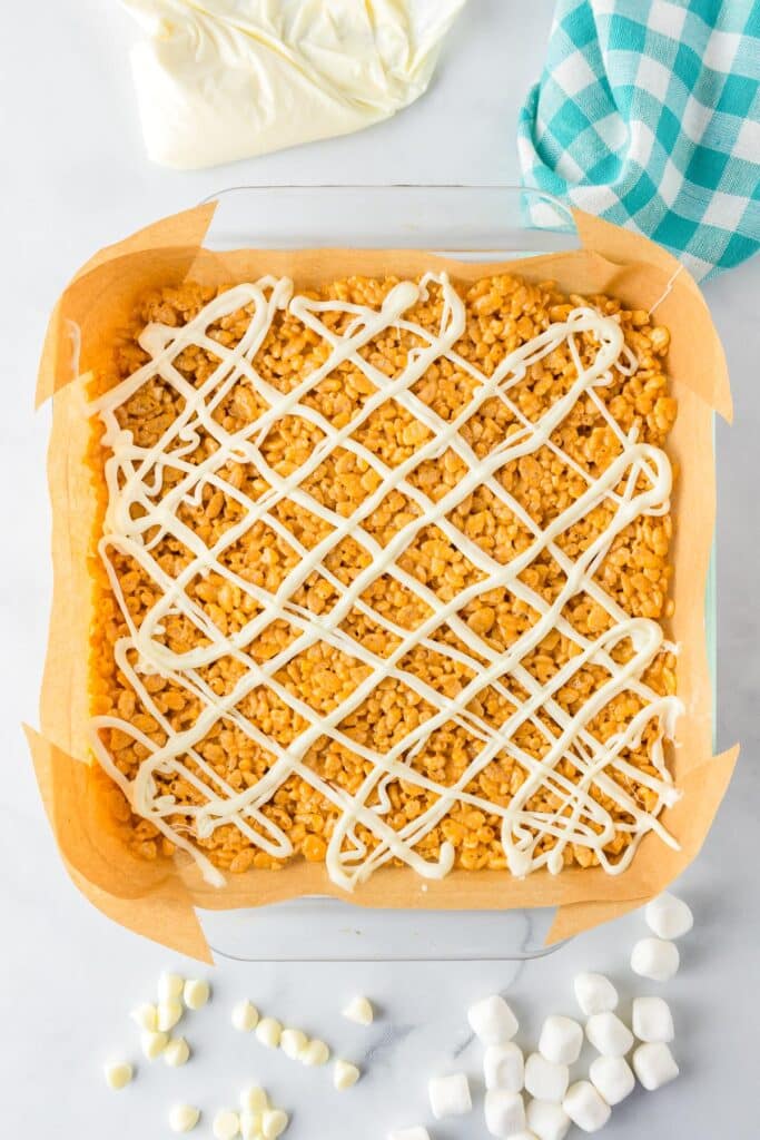 A square baking dish lined with parchment paper, filled with pumpkin spice rice crispy treats drizzled with white chocolate, with a piping bag of melted white chocolate nearby.