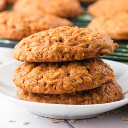 Square view of three oatmeal pumpkin cookies stacked on a white plate with more cookies on a wire rack behind.