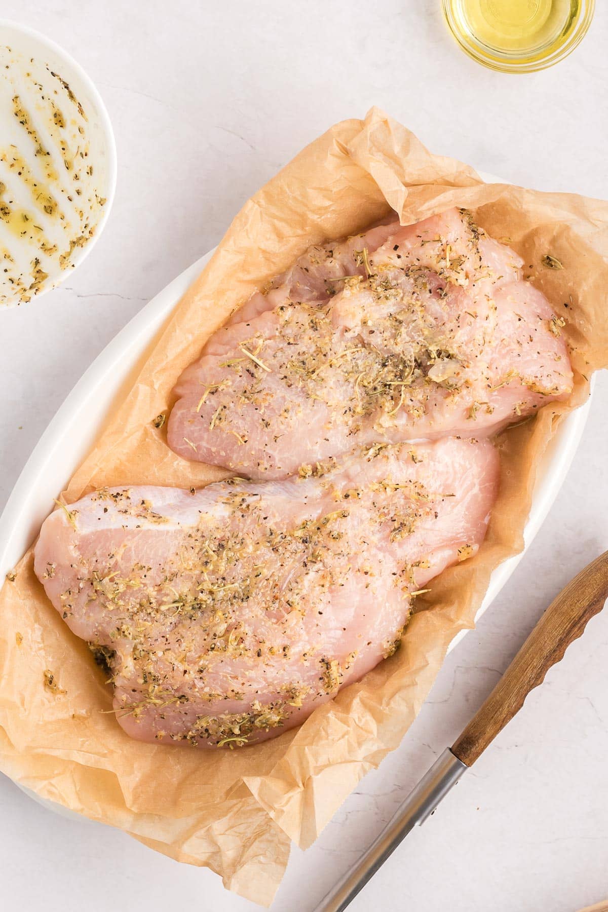 Two pieces of raw, turkey tenderloin placed on parchment paper in an oval dish seasoned with a butter and herb mixture.