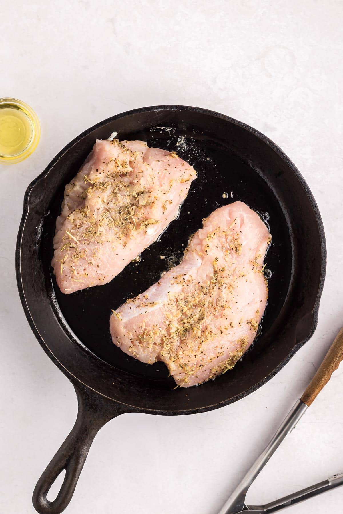 Two raw, seasoned turkey tenderloins in a black cast iron next to tongs.