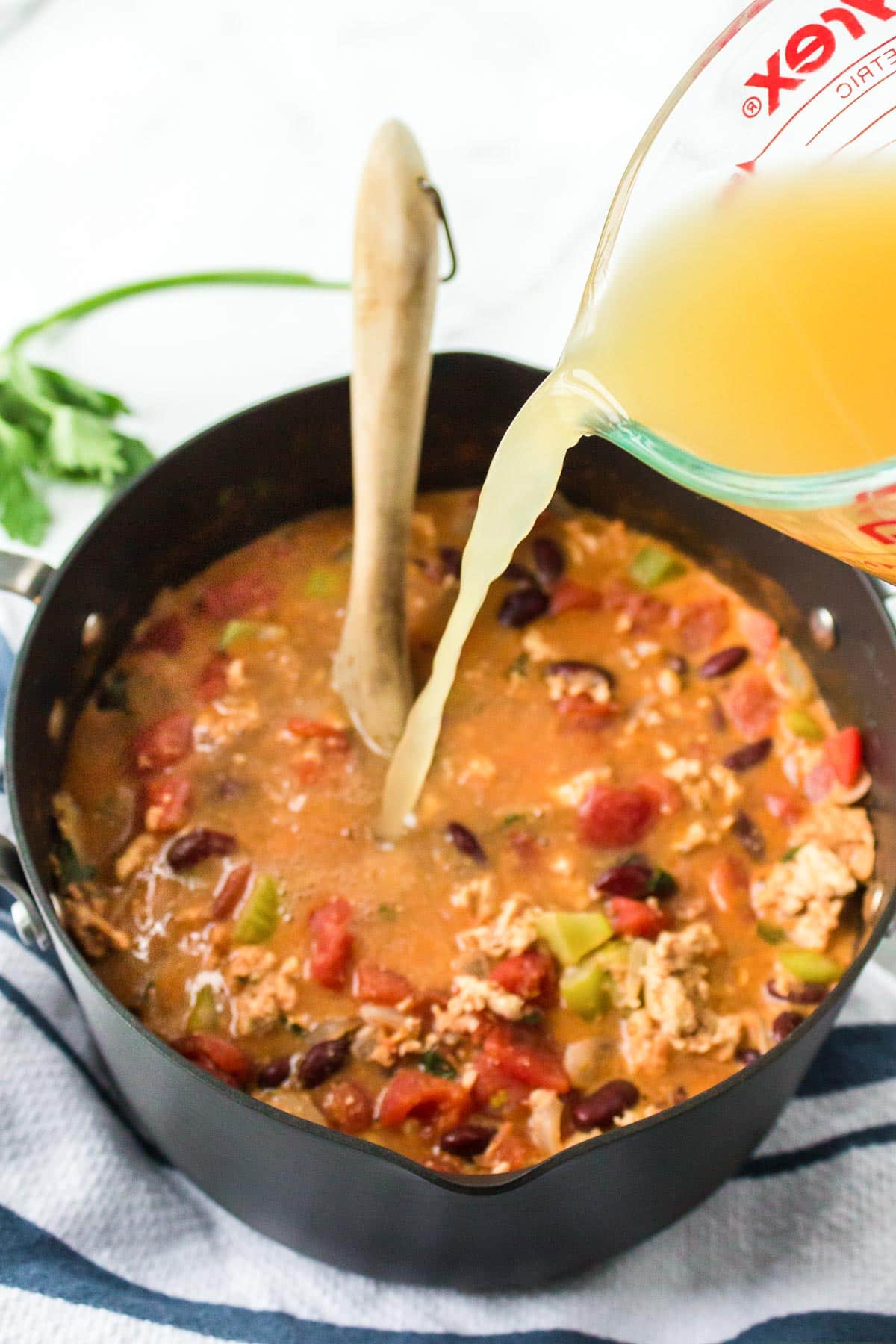 A measuring cup pouring chicken broth into a pot of turkey chili.