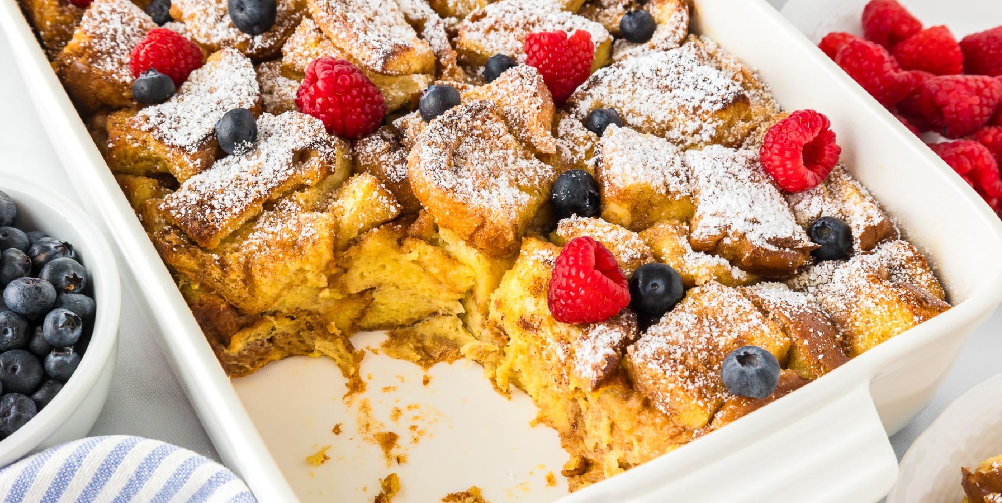 A wide view of a brioche french toast casserole topped with berries and powdered sugar in a baking pan.