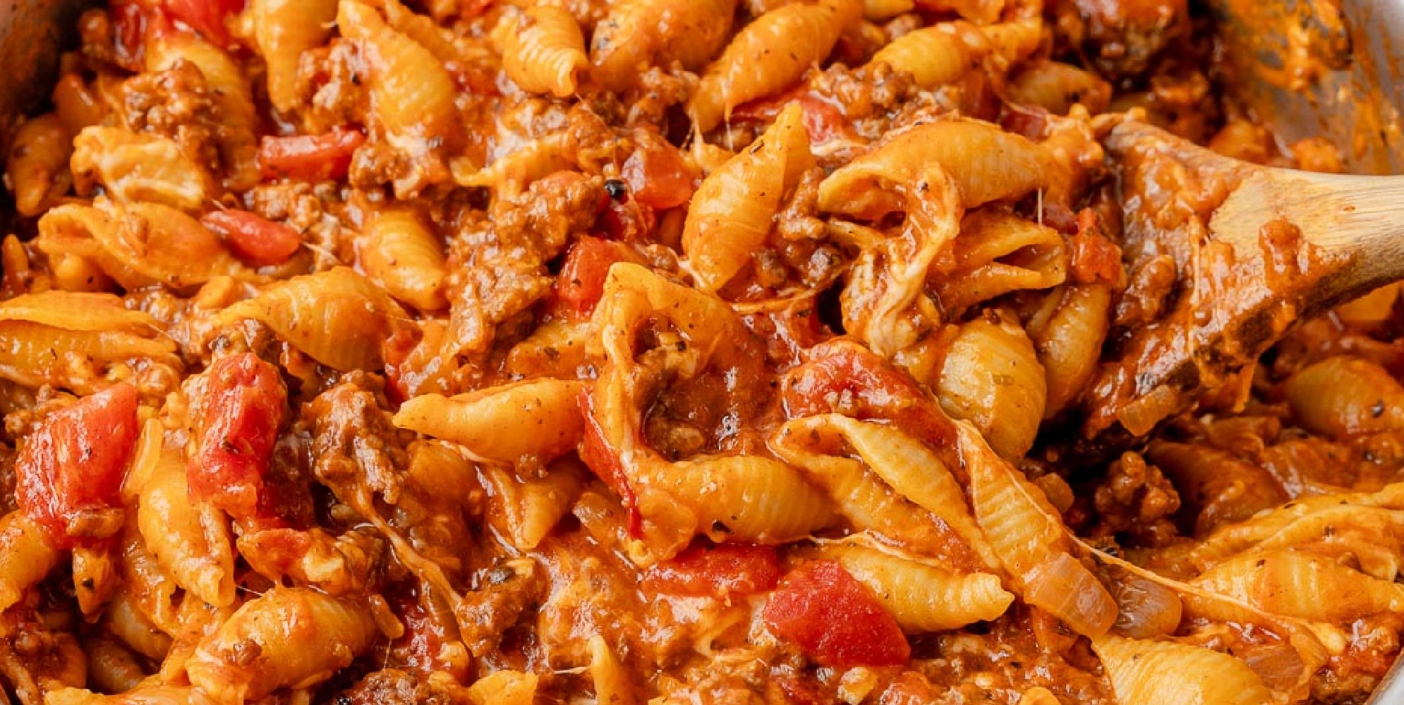 A wide view of a wooden spoon stirring a skillet filled with cheesy homemade hamburger helper in a pan.