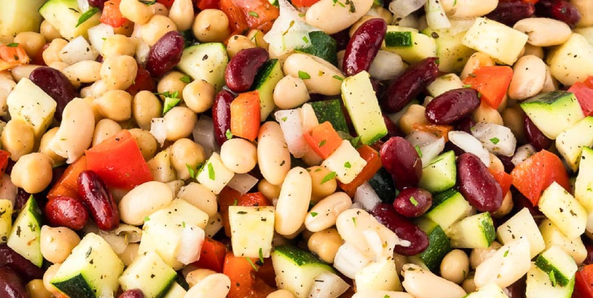 A close-up of a mixed bean salad with chickpeas, kidney beans, white beans, diced zucchini, red bell peppers, and chopped herbs.