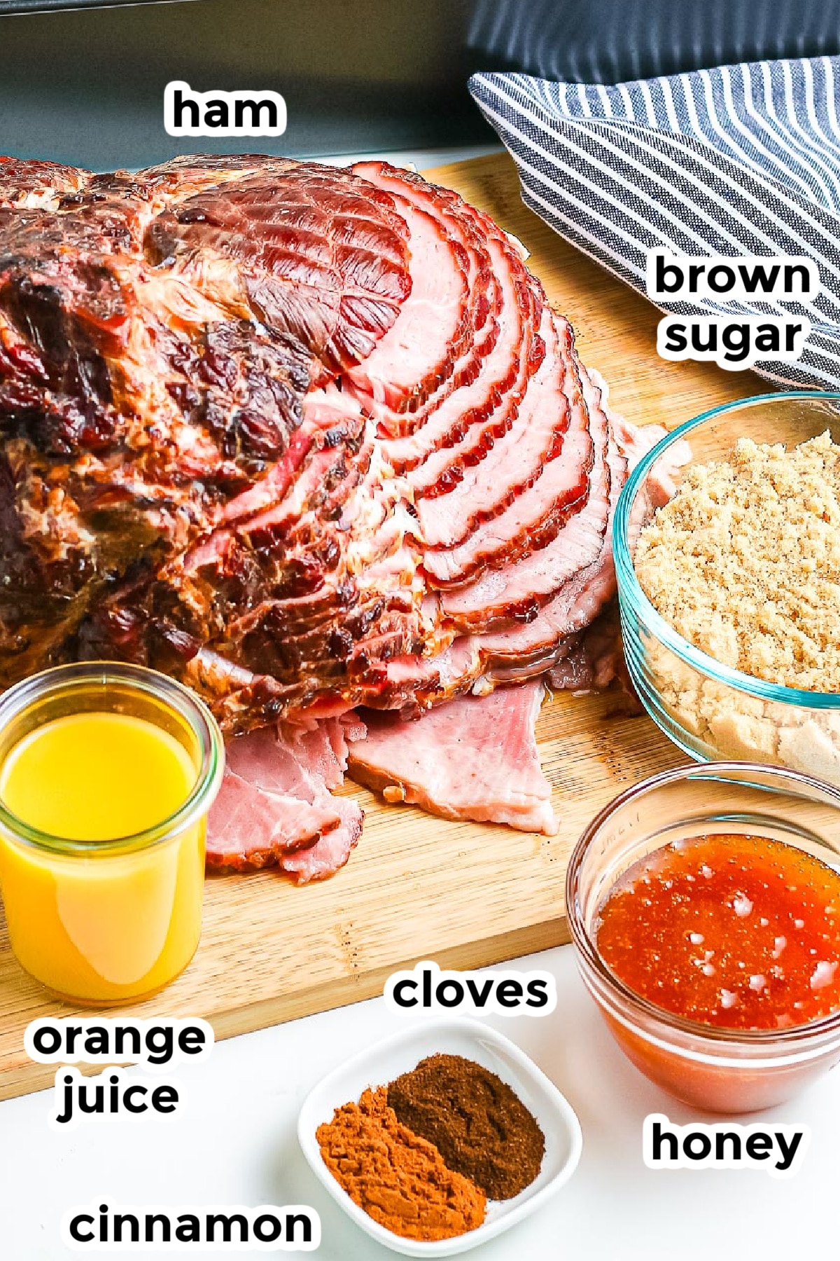 Sliced ham on a wooden board with labeled ingredients for ham glaze around it in bowls including orange juice, brown sugar, honey, cinnamon and cloves.