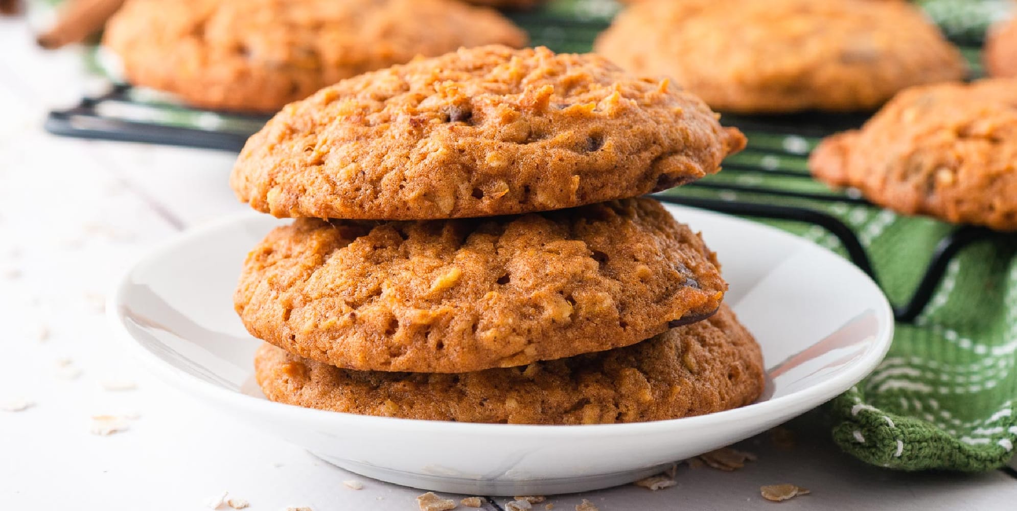 Wide view of three oatmeal pumpkin cookies stacked on a plate with a towel and more cookies on a wire rack in the background.