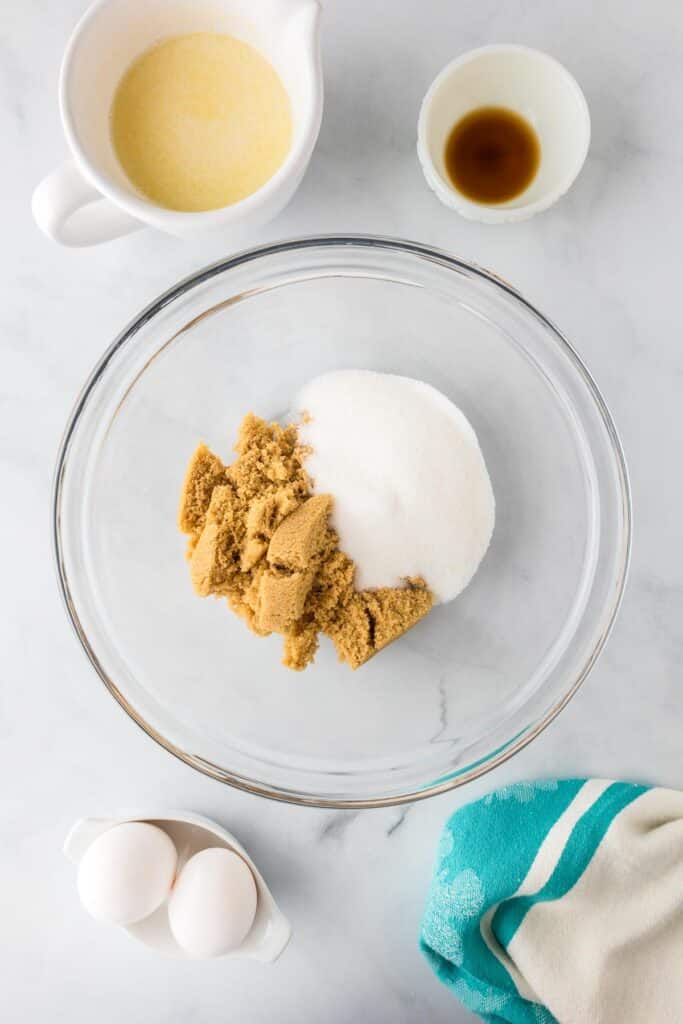 A glass bowl contains white and brown sugar for Andes mint brownies with eggs, melted butter and vanilla in bowls nearby to mix the wet part of the batter.