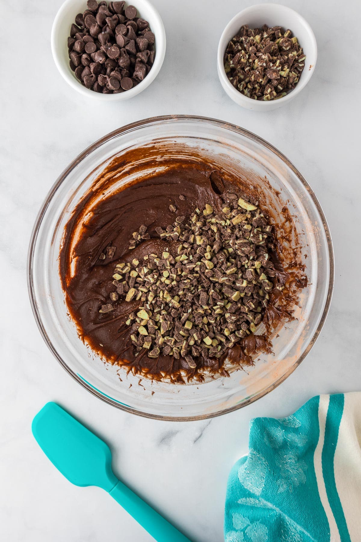 A glass bowl containing chocolate andes mint brownie batter being mixed with crushed andes mint pieces with chocolate chips and more andes mint pieces in bowls nearby on the counter.