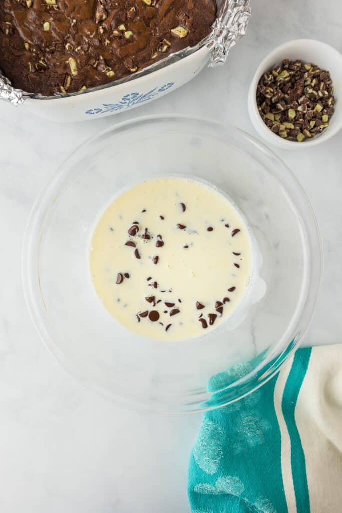 A mixing bowl with cream and chocolate chips with andes mint pieces and a pan of baked Andes mint brownies nearby.