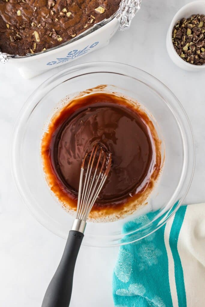 A whisk in a bowl of melted chocolate sits next to a pan of Andes Mint brownies.